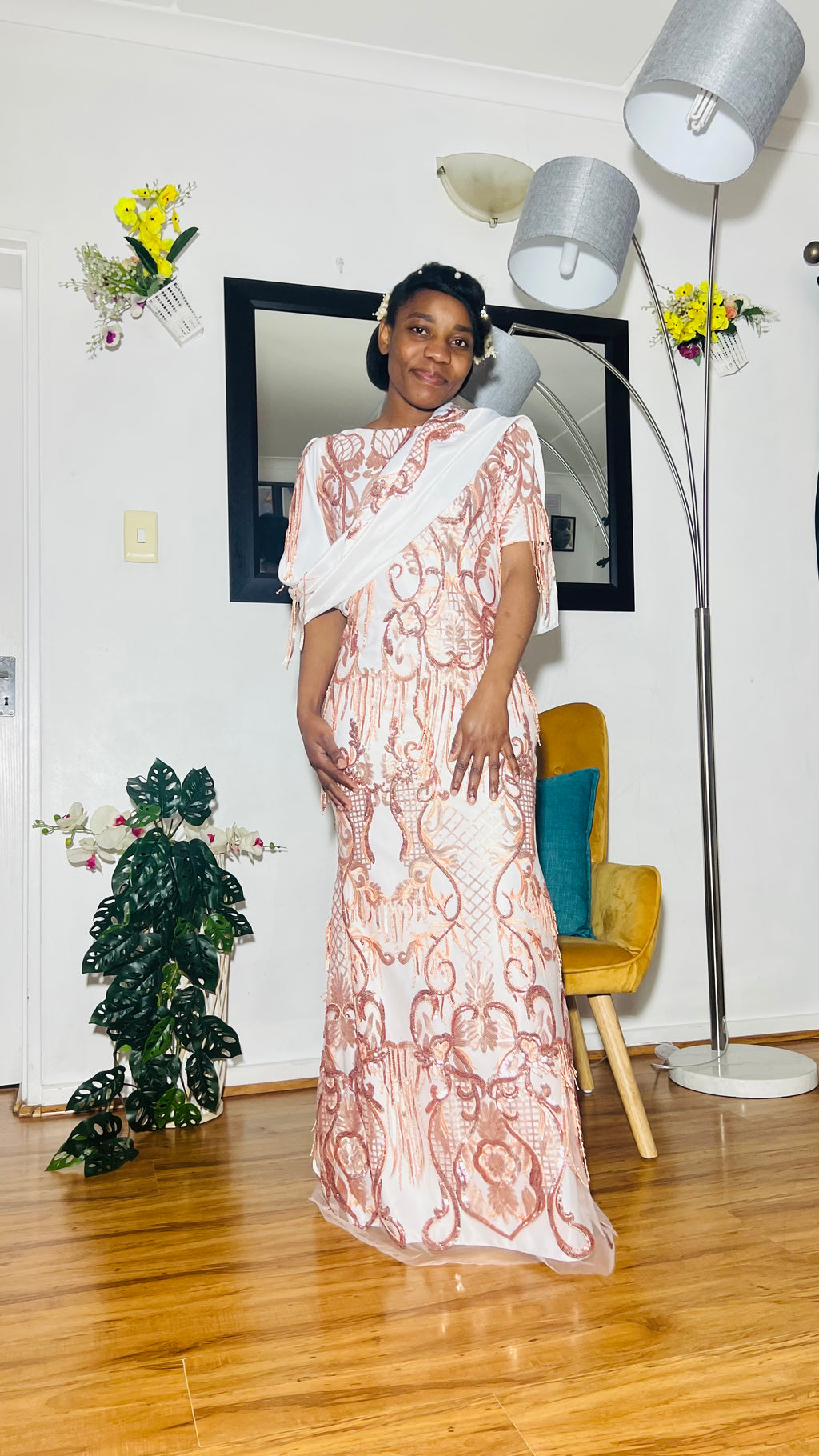 Woman in a white lace dress standing in a room with a lamp and chair.